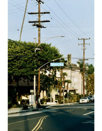 A vintage-style street scene photograph showing a road corner with utility poles, trees, a blue street sign reading 'Beverly Ave', houses, parked cars, and a bicycle symbol painted on the pavement.