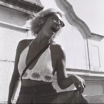 A black and white fashion photograph of a woman laughing outdoors, wearing a white crochet halter crop top and dark sunglasses, with a building facade in the background.