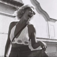 A black and white fashion photograph of a woman laughing outdoors, wearing a white crochet halter crop top and dark sunglasses, with a building facade in the background.