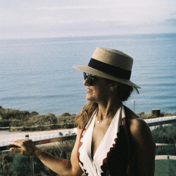 A woman in profile wearing a straw boater hat with a black band, dark sunglasses, and a white halter top with scalloped edges, standing on a balcony with the ocean in the background.