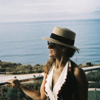 A woman in profile wearing a straw boater hat with a black band, dark sunglasses, and a white halter top with scalloped edges, standing on a balcony with the ocean in the background.