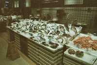 A market seafood display with fish and shellfish on ice, shrimp, and other seafood in a real indoor market setting with tiled walls and signage.