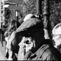 A black and white street photograph featuring an elderly man in the foreground wearing a flat cap, sunglasses, and a jacket, with other pedestrians and city buildings blurred in the background.