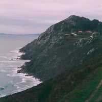 A scenic coastal landscape with a steep rocky cliff rising above the ocean, white surf along the shoreline, and a grassy path in the foreground under a pale overcast sky.