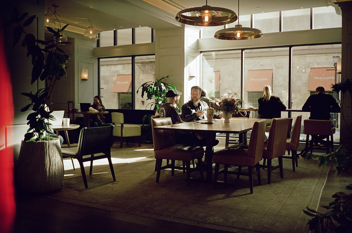A dimly lit modern dining room or cafe interior with several people seated around a round wooden table, large windows, hanging pendant lights, and potted plants.