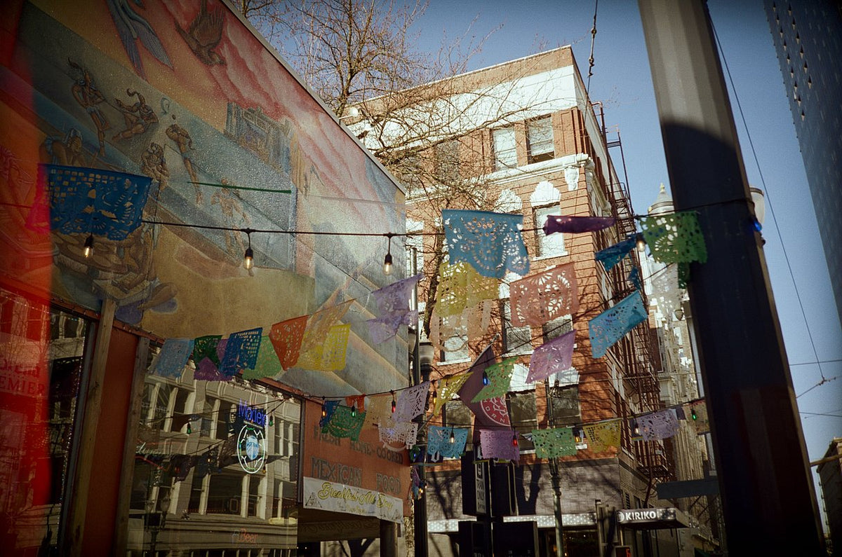 A narrow urban street lined with colorful hanging papel picado banners and a large mural on the left building, with brick buildings and bare trees in the background under a blue sky.
