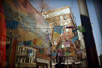 A narrow urban street lined with colorful hanging papel picado banners and a large mural on the left building, with brick buildings and bare trees in the background under a blue sky.