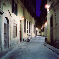 A narrow cobblestone street at night between old buildings, with warm wall lights glowing, a small scooter parked near the center, and a deep blue-purple sky above.