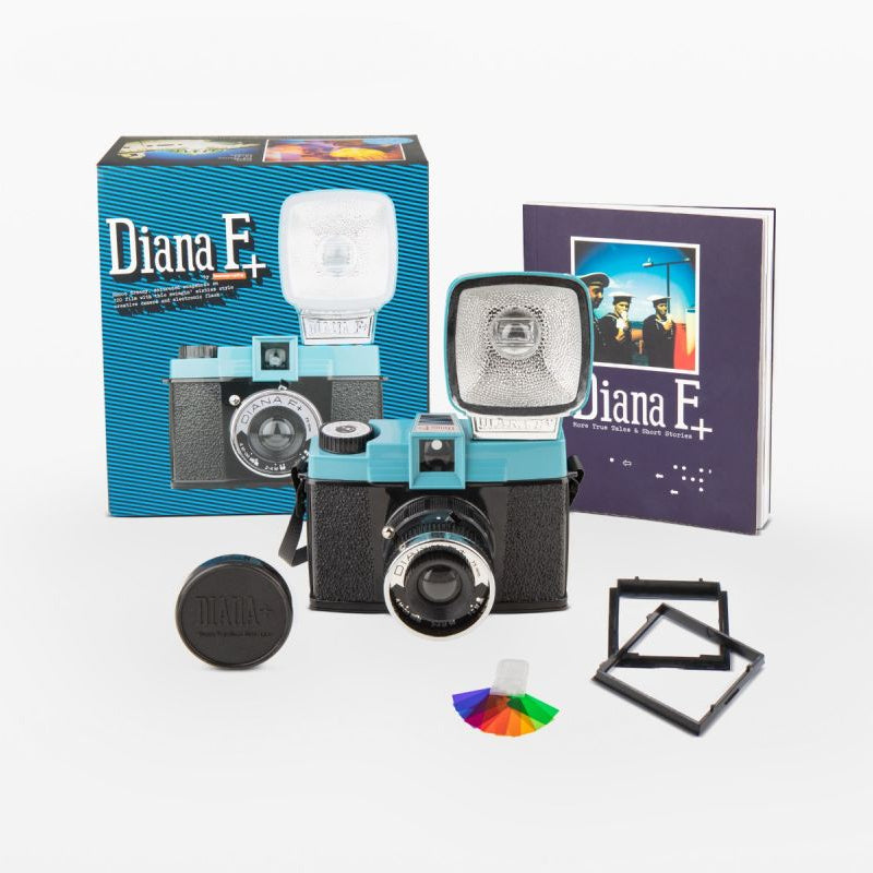 Diana F+ color camera kit with a turquoise and black camera body, a light speedlight, original box, instruction booklet, lens cap, filter, and frame on a white background.