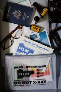 A white mesh zippered pouch labeled 'FILM DO NOT X-RAY' sits on a white surface surrounded by travel-themed props including a passport, film canister, camera strap, glasses, and printed cards.