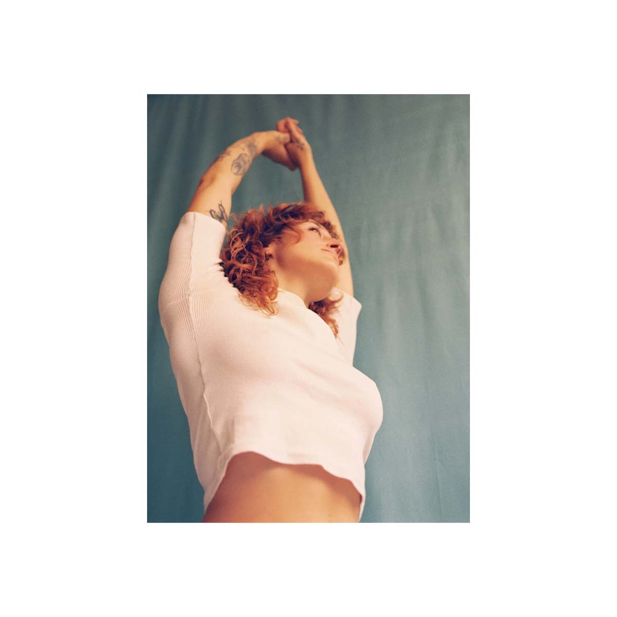 A person with curly red hair wearing a white cropped long-sleeve top, looking upward with arms raised, set against a muted blue-green studio backdrop.