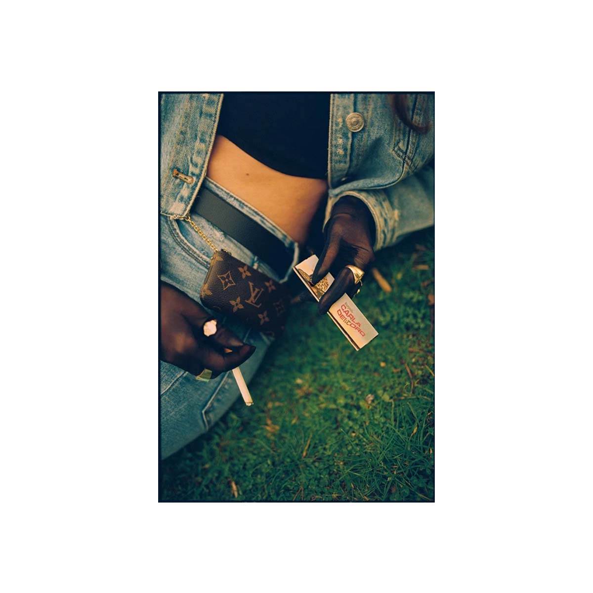 A person wearing a light-wash denim jacket and jeans sits on grass, holding a cigarette and a small Louis Vuitton monogram wallet. A cigarette pack is also visible.