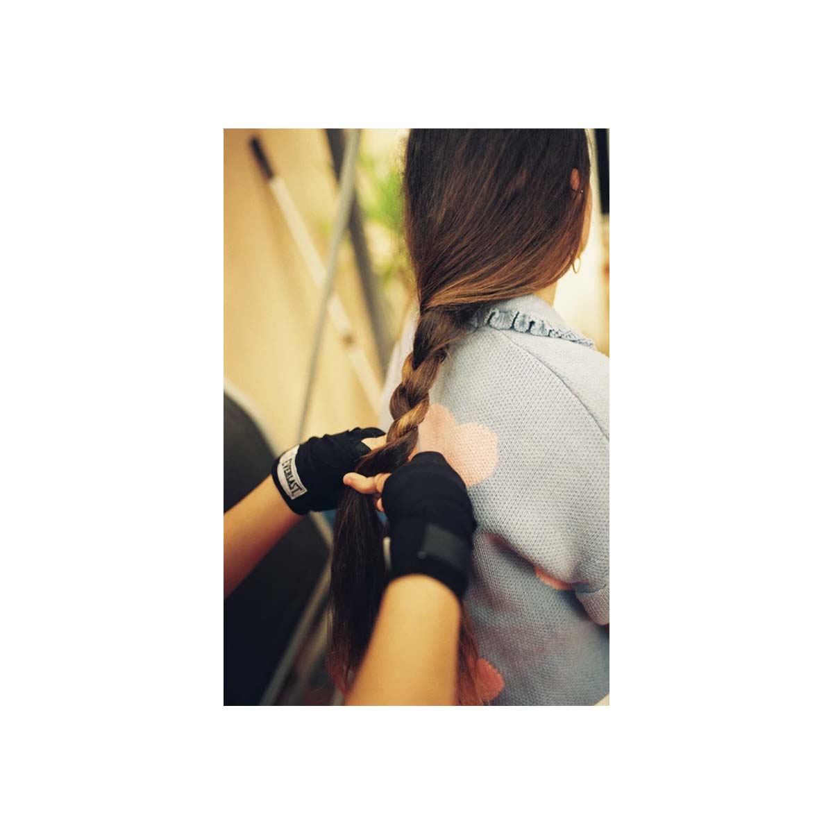 A person with long brown hair seen from behind, having their hair braided by another person wearing black fingerless gloves. The person wearing the product is dressed in a light gray top, and the background is a softly blurred indoor setting.