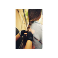 A person with long brown hair seen from behind, having their hair braided by another person wearing black fingerless gloves. The person wearing the product is dressed in a light gray top, and the background is a softly blurred indoor setting.