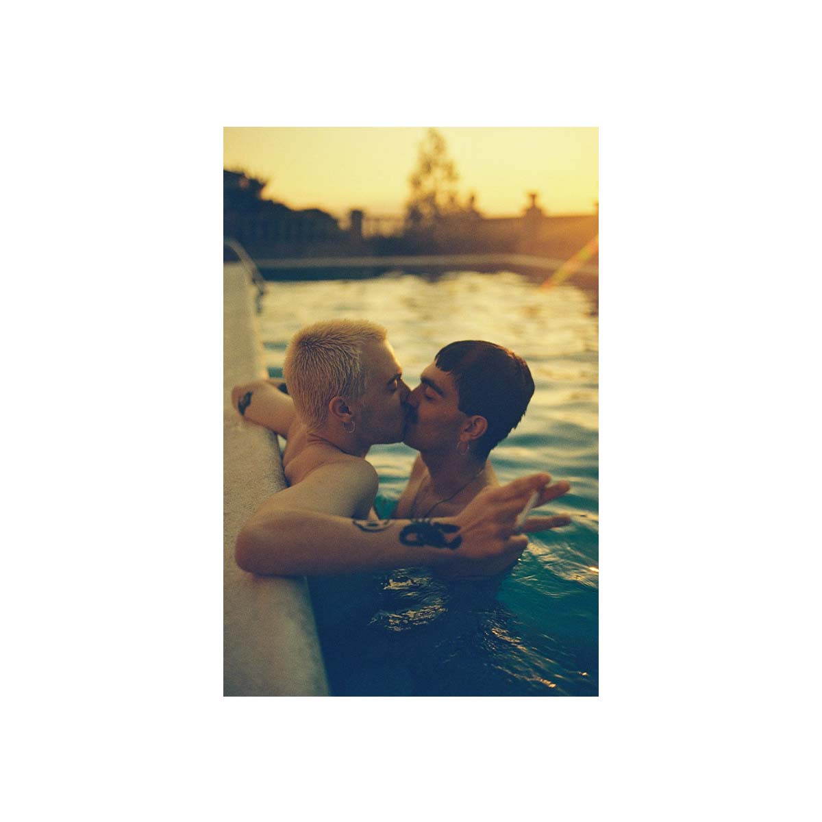 A vertical photographic print showing two people kissing in a pool at sunset, centered on a white background.