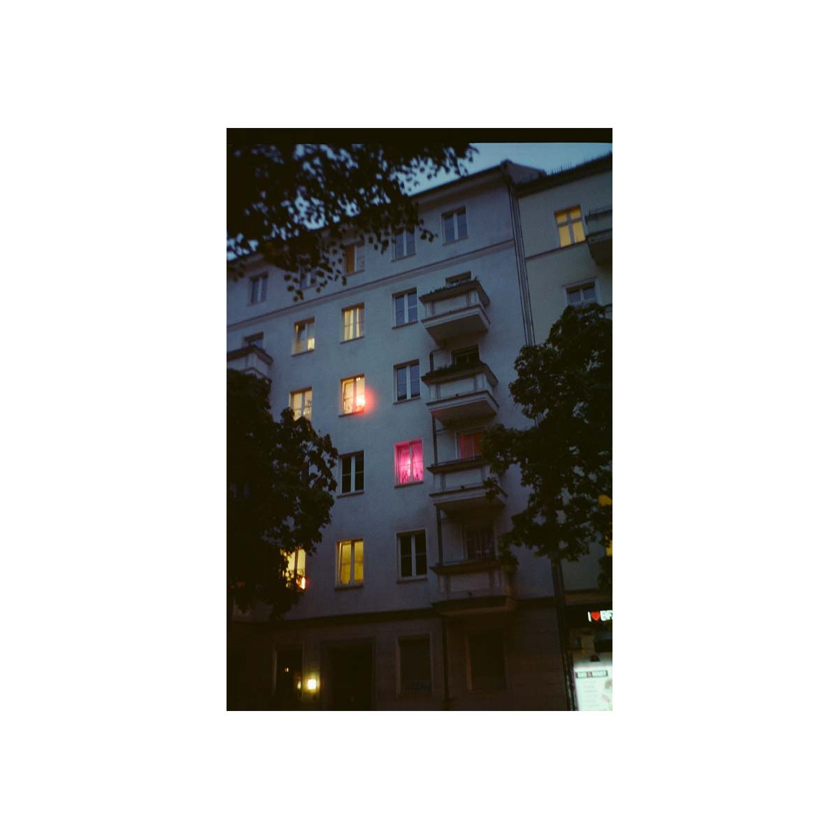 A vertical photographic print of a European apartment building at dusk, framed by a wide white border. The building has a light gray facade with balconies and several windows lit with warm yellow light, including one glowing with a red-pink hue.