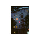 A vertical photographic print of a European apartment building at dusk, framed by a wide white border. The building has a light gray facade with balconies and several windows lit with warm yellow light, including one glowing with a red-pink hue.