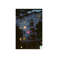 A vertical photographic print of a European apartment building at dusk, framed by a wide white border. The building has a light gray facade with balconies and several windows lit with warm yellow light, including one glowing with a red-pink hue.