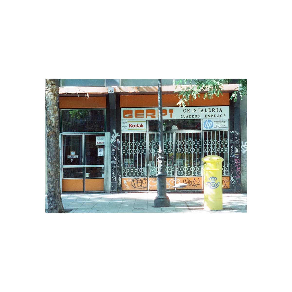 A street scene photograph of a storefront with orange and white signage reading 'Cristalería Cajas Ríos' and a visible Kodak logo, with metal security gates, sidewalk, tree trunk, lamp post, and a yellow trash can.