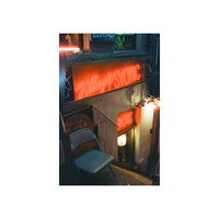 A vertical photographic print of a dimly lit alleyway at night, featuring a glowing orange-red neon sign that reads 'Village Psychic' above a brick wall and doorway, with a folding chair in the foreground.