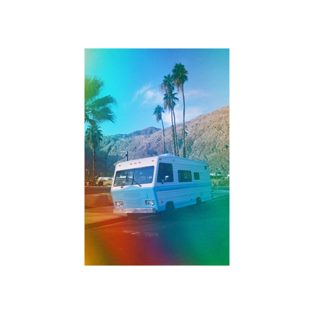 A vintage white motorhome is parked on a road with palm trees and mountains in the background under a bright blue sky, with a colorful rainbow lens flare effect around the edges.