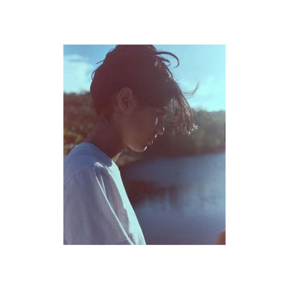 A square photo print featuring a side-profile portrait of a young man with dark hair, wearing a white t-shirt, set against a bright blue sky and water background, presented on a white border.