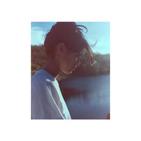 A square photo print featuring a side-profile portrait of a young man with dark hair, wearing a white t-shirt, set against a bright blue sky and water background, presented on a white border.