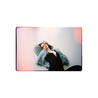 A square photographic print or poster featuring a woman reclining against a pale background with pink and orange light leaks, shown centered on a plain white background.