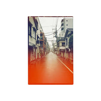 A vertical photographic print of a narrow urban street lined with buildings, shown centered on a plain white background. The print has a warm orange-red border and a muted, slightly vintage-toned street scene inside.