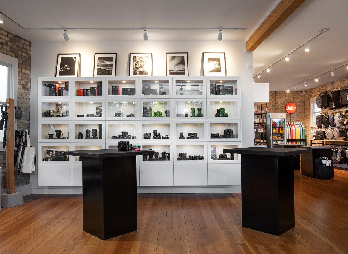 A photography equipment store interior with a large white shelving unit displaying cameras, lenses, and accessories, with two black display pedestals in the foreground on a wood floor.