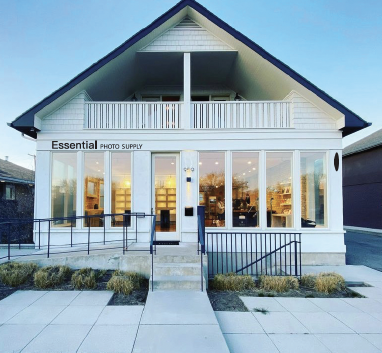 Front exterior of a white two-story building with large glass windows and a sign reading 'Essential Photo Supply' on the left side of the facade.