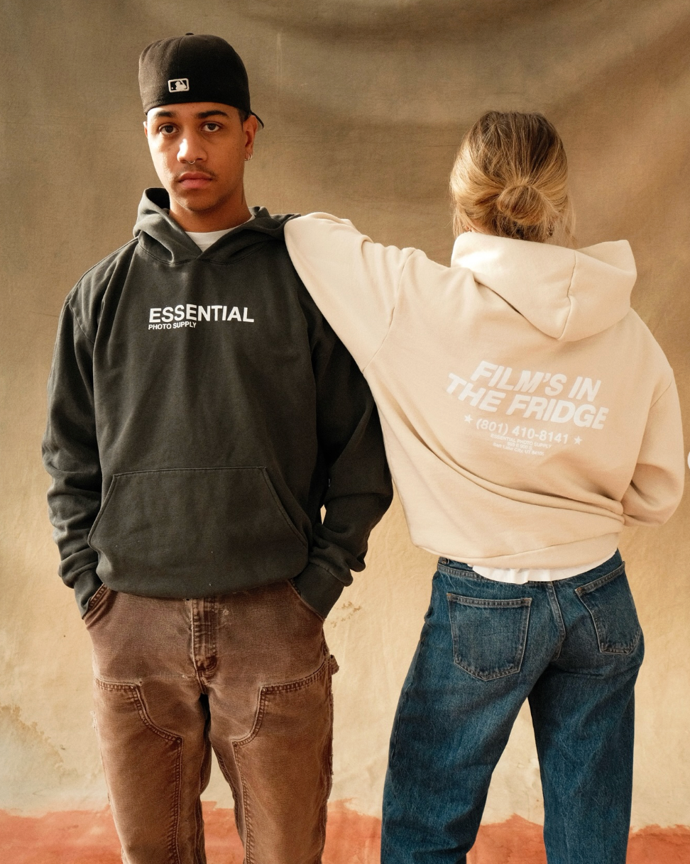 Two people wearing hoodies with text, one black and one beige, on a neutral background.