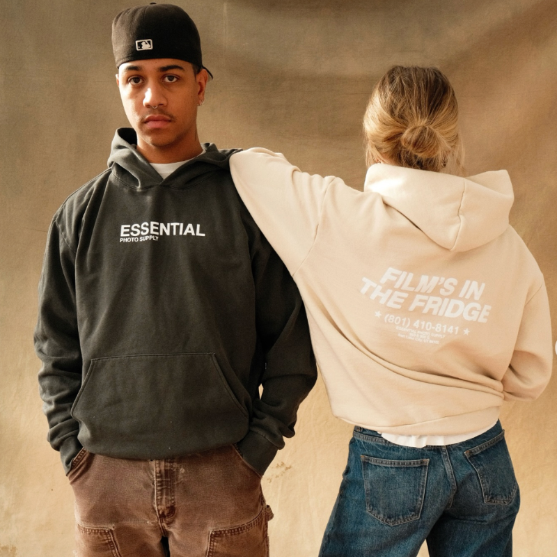 Two people wearing hoodies with text, one black and one beige, on a neutral background.