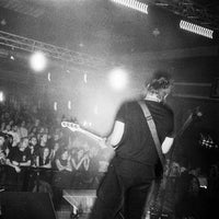 Black and white photograph of a live music performance on stage, showing a guitarist from behind facing a large crowd in a concert hall.