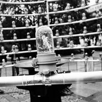 A black and white photograph of a heavy-duty industrial warning beacon light mounted on a metal pipe, with scaffolding and a crowd of people visible in the blurred background.