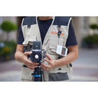 A man wearing a beige and navy Godox multi-pocket utility vest holds a camera with a zoom lens mounted on a tripod. A white Godox remote is clipped to his left chest pocket, and the Godox logo is printed on the right chest pocket.