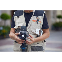A man wearing a beige and navy Godox multi-pocket utility vest holds a camera with a zoom lens mounted on a tripod. A white Godox remote is clipped to his left chest pocket, and the Godox logo is printed on the right chest pocket.