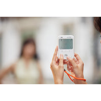 A person holding a white Godox handheld light meter with an orange wrist strap, shown in a real-world blurred environment.