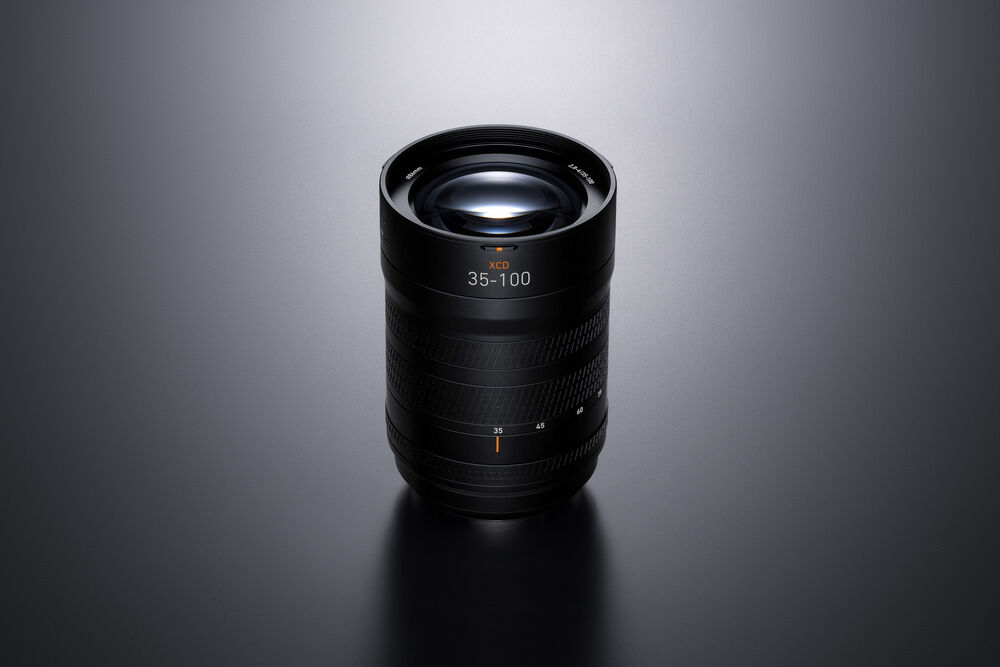 A black camera lens with '35-100' printed on the front, photographed on a dark gray reflective surface with a soft spotlight from above.