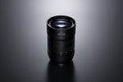 A black camera lens with '35-100' printed on the front, photographed on a dark gray reflective surface with a soft spotlight from above.