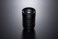 A black camera lens with '35-100' printed on the front, photographed on a dark gray reflective surface with a soft spotlight from above.