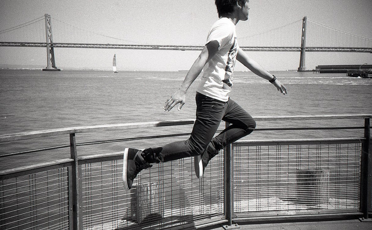 A black and white lifestyle photograph of a person mid-jump over a metal railing, with a large suspension bridge and water in the background.