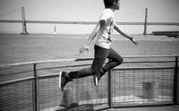 A black and white lifestyle photograph of a person mid-jump over a metal railing, with a large suspension bridge and water in the background.