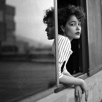 Black and white portrait of a woman with curly hair leaning against a window frame, wearing a vertically striped top. The photo has a reflective glass surface on the left side.