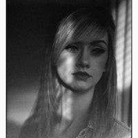 A black and white portrait photograph of a young woman with long hair, wearing a light denim shirt, framed with a thin black border and a small photographer credit at the bottom right.
