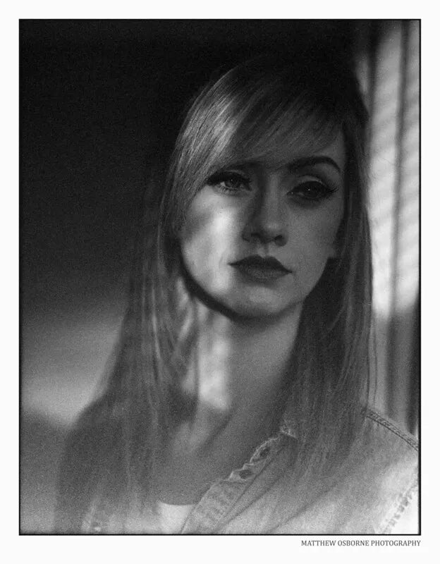 A black and white portrait photograph of a young woman with long hair, wearing a light denim shirt, framed with a thin black border and a small photographer credit at the bottom right.