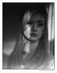 A black and white portrait photograph of a young woman with long hair, wearing a light denim shirt, framed with a thin black border and a small photographer credit at the bottom right.