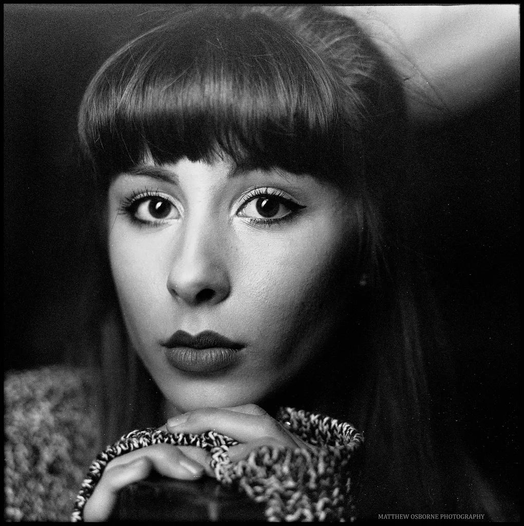 Black and white portrait photograph of a woman with long dark hair, straight bangs, and a textured knit top, photographed in a moody studio setting.
