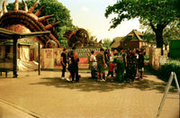 A vintage photograph of a crowd of people standing in front of a large, whimsical dragon-themed entrance at what appears to be a fair or amusement venue. The scene has a strong warm, yellowed, and grainy aesthetic typical of older film or scan scans.