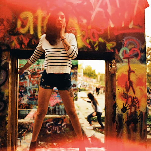 A woman wearing a white long-sleeve striped crop top and black denim shorts stands in front of a graffiti-covered wooden frame, with a blurred outdoor street scene behind her.